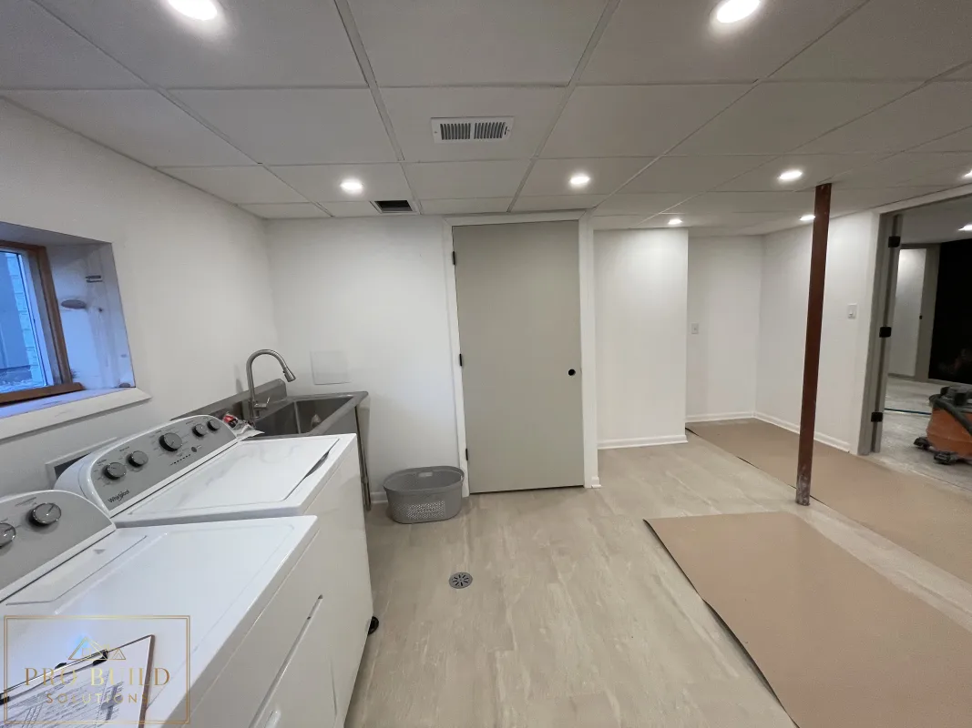 Views of the New Storage Door in the Basement finishing project with laundry space, stainless steel sink, recessed ceiling lights, closet doors, and durable plank flooring before Final Paint, Western Springs