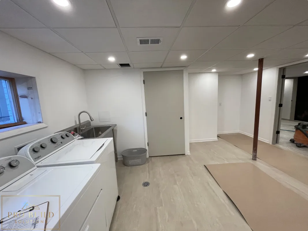 Views of the New Storage Door in the Basement finishing project with laundry space, stainless steel sink, recessed ceiling lights, closet doors, and durable plank flooring before Final Paint, Western Springs
