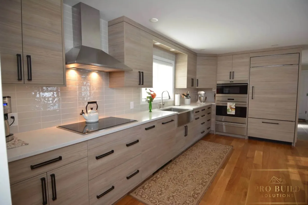 Contemporary galley kitchen featuring natural wood cabinetry, white counters, glossy tile backsplash, and black hardware, Barrington